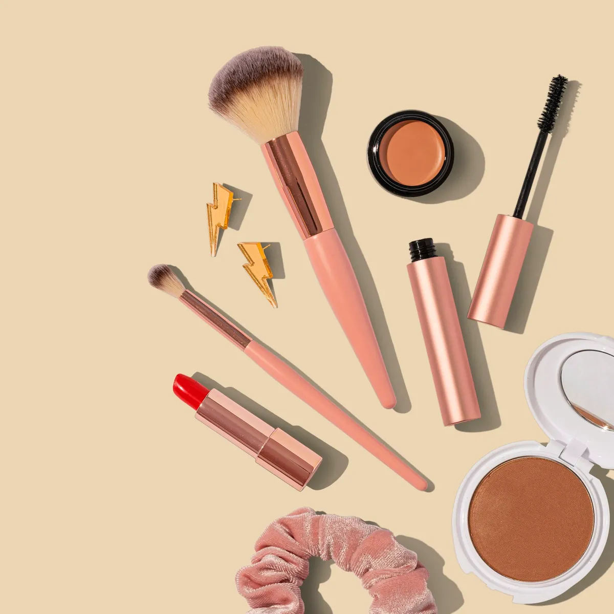 Brochas de maquillaje, máscara de pestañas, pintalabios y polvos compactos sobre fondo crema